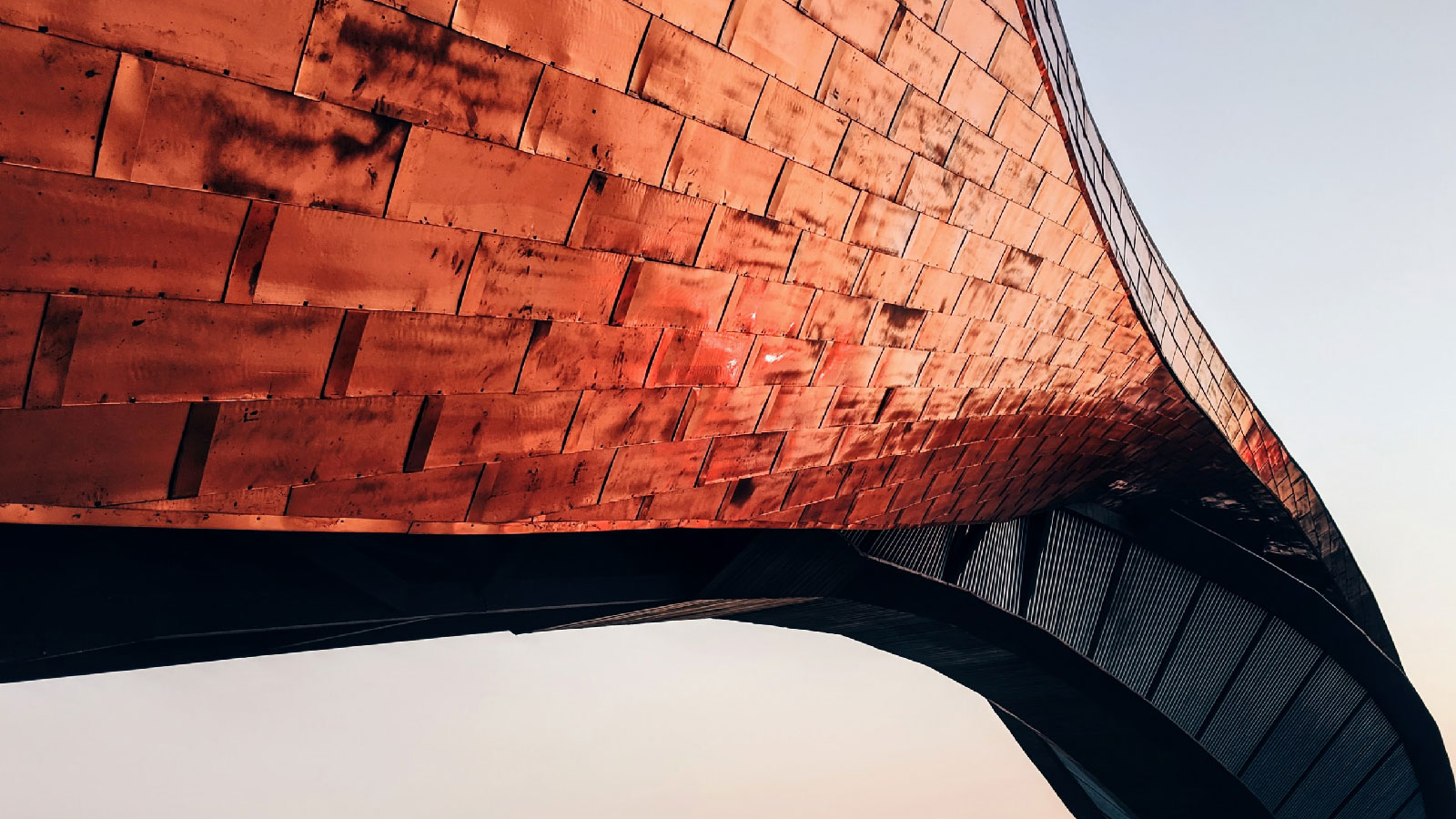 Low-angle, close-up abstract view of a building facade covered in overlapping copper-colored rectangular metal panels.