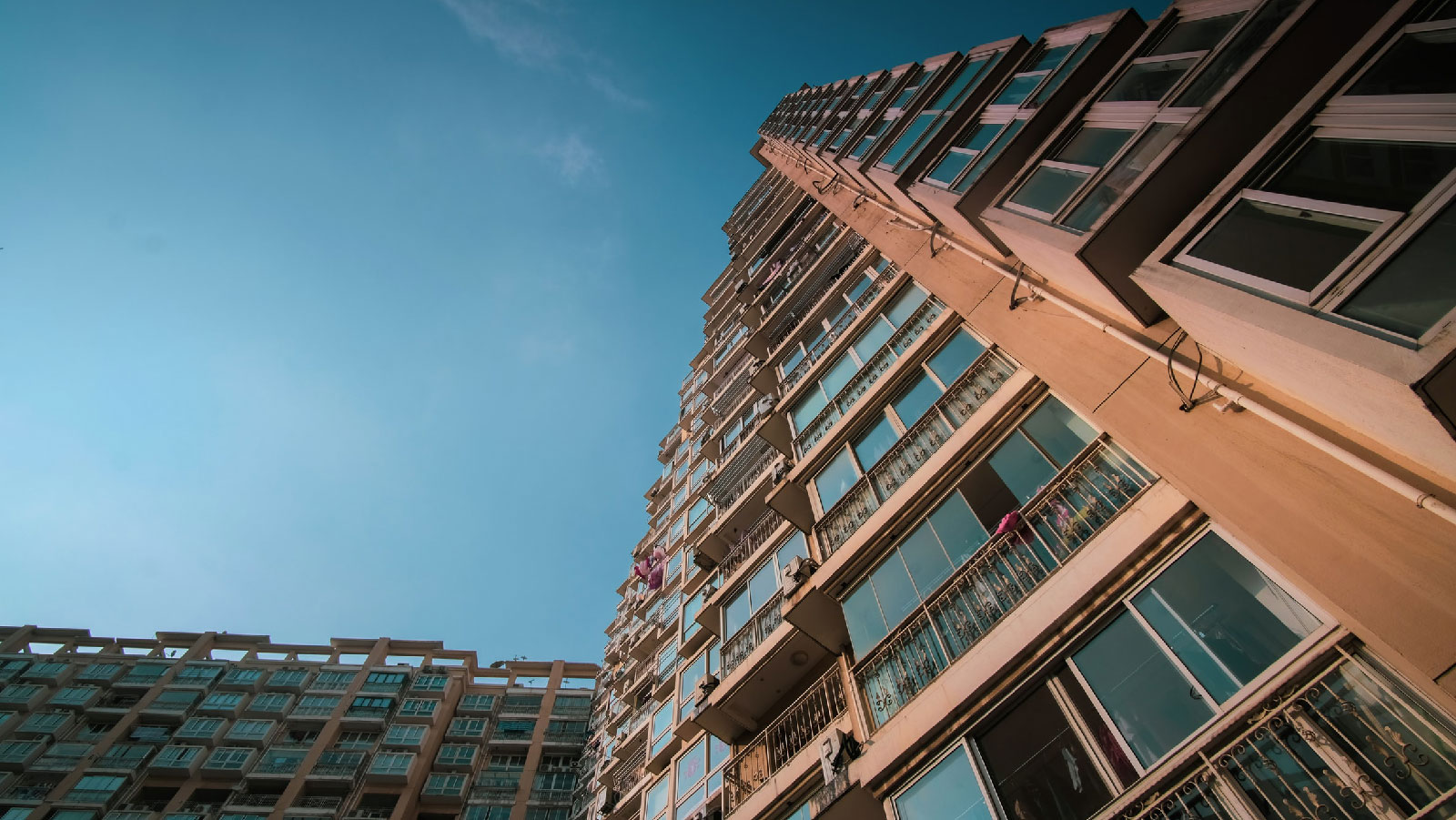 Low-angle view of a tall, beige residential apartment building with many windows against a clear blue sky.