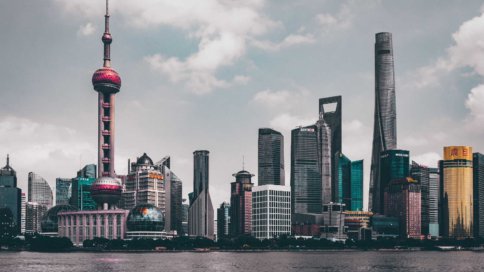 Panoramic view of the Shanghai city skyline, including the Oriental Pearl Tower, across the water under a cloudy sky.
