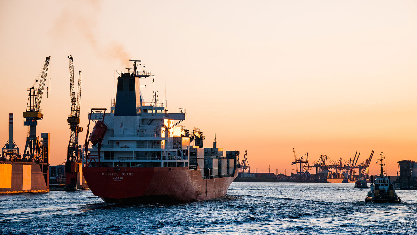 A red container ship named 'CHARLES ISLAND NASSAU' leaves a bustling port at sunset, with numerous cranes in the background.