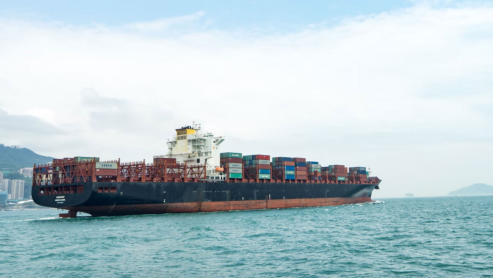 A massive container ship loaded with cargo sails on the ocean near a coastal city under a cloudy sky.