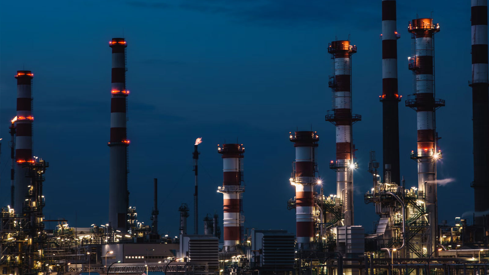 Oil refinery or chemical plant at night, with illuminated industrial towers, smokestacks, and a flare.