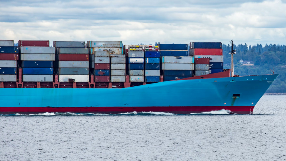 Close-up view of the bow of a large, bright blue container ship sailing on the sea, loaded with multicolored cargo.