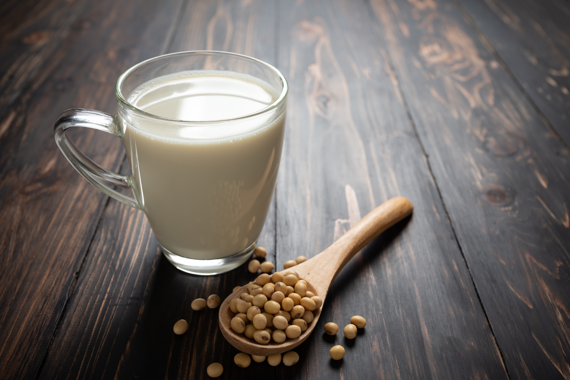 A glass mug of light-colored soy milk next to a wooden spoon filled with raw soybeans on a dark wood table.