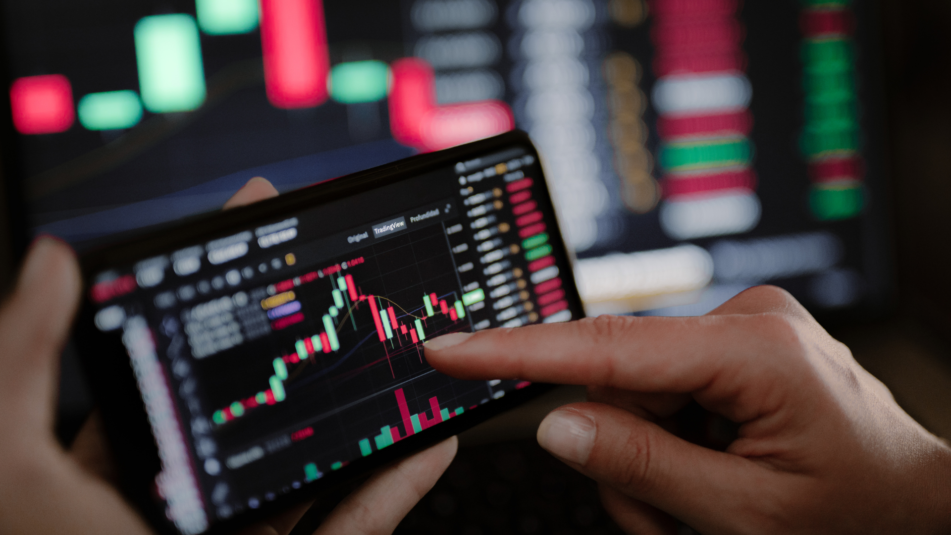A finger tapping a candlestick chart on a horizontal smartphone screen, with a blurry background of a larger monitor display.