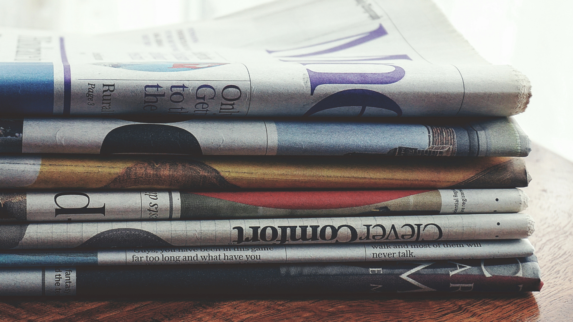 A neatly stacked pile of folded newspapers resting on a wooden surface, showing various headlines and print media layers.
