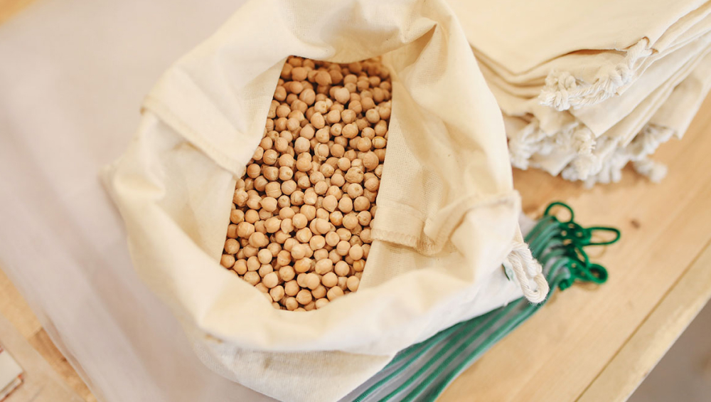 Overhead view of a tan cloth bag filled with dry, raw chickpeas (garbanzo beans) on a wooden table next to reusable cloth sacks.