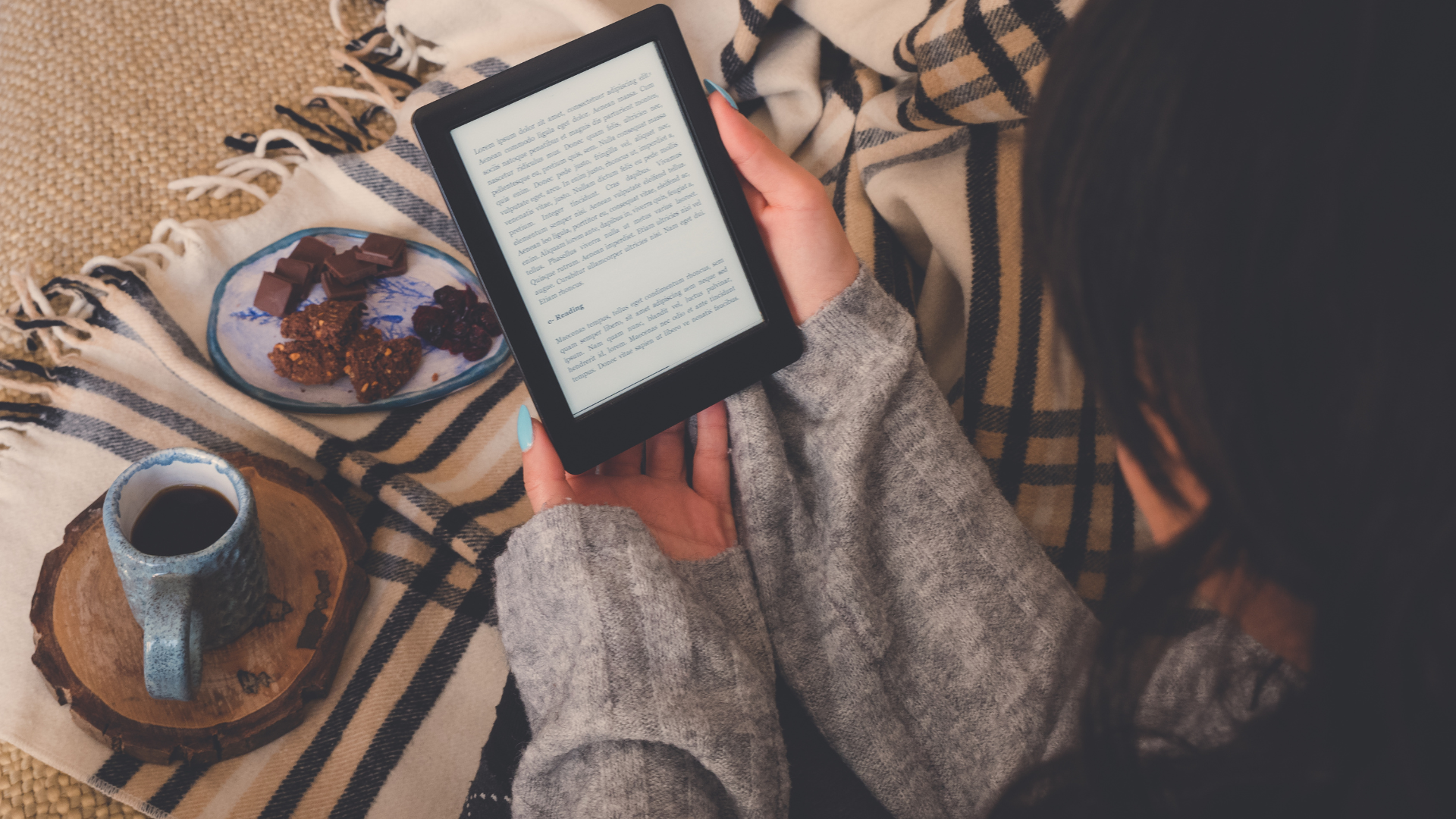 Overhead shot of a person relaxing with a cozy blanket, reading an e-reader (e-book) while enjoying coffee and snacks (chocolate/berries).
