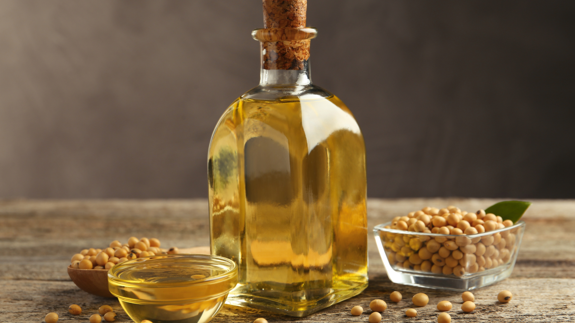 Clear bottle and small dish of golden soybean oil surrounded by raw soybeans on a rustic wooden table.