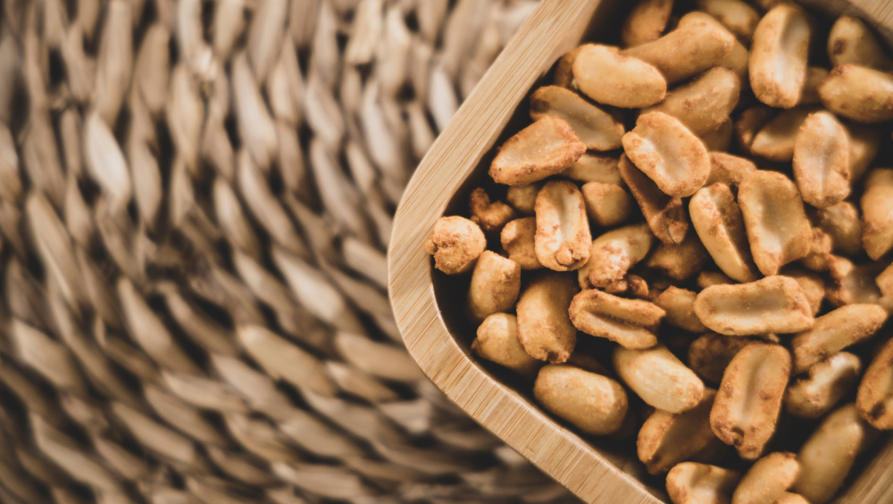 Close-up of roasted and salted peanuts in a small wooden bowl, resting on a woven, wicker-like surface.