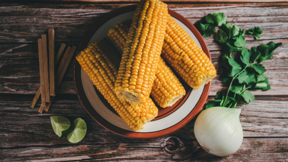 Boiled corn on the cob served on a brown plate with an onion, lime wedges, and cilantro on a wooden table.