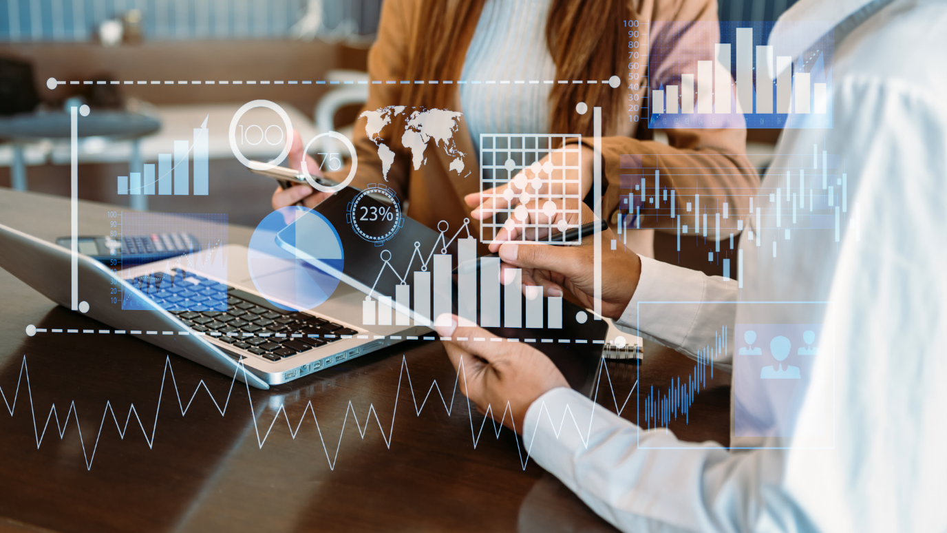 Two business people discussing data on a laptop, with multiple holographic financial charts, graphs, and a world map overlaid.