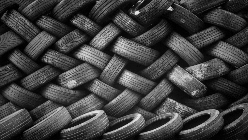 A densely stacked pattern of black, used vehicle tires forming a wall or woven structure, viewed in black and white.