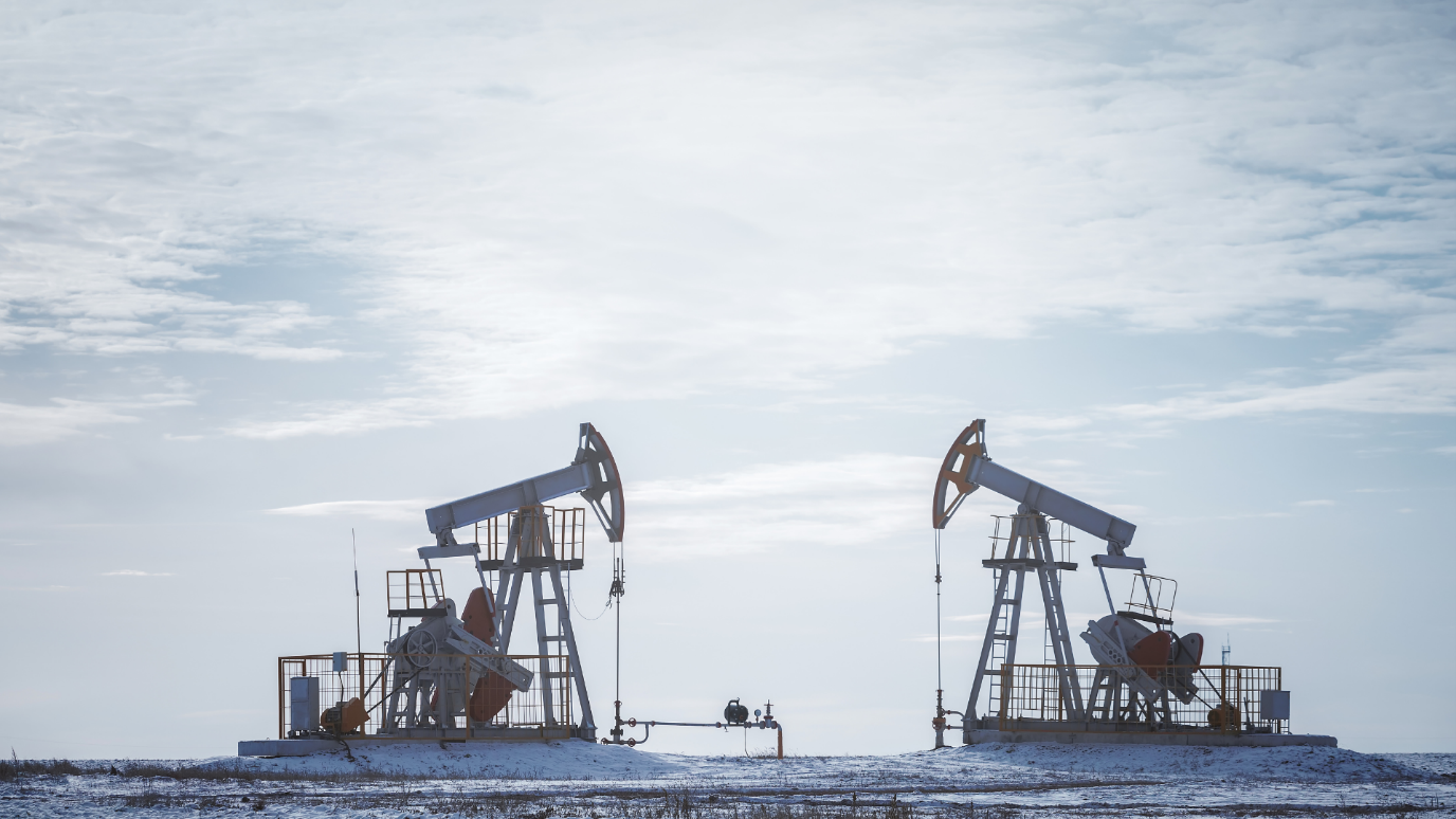 Two silhouetted oil pumpjacks (nodding donkeys) operating in a vast, flat, snowy field under a cloudy sky.
