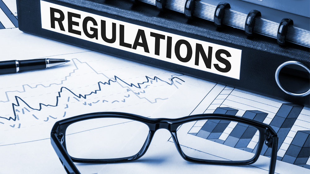 A black ring binder labeled "REGULATIONS" lying on a desk with financial charts, graphs, a pen, and a pair of reading glasses.