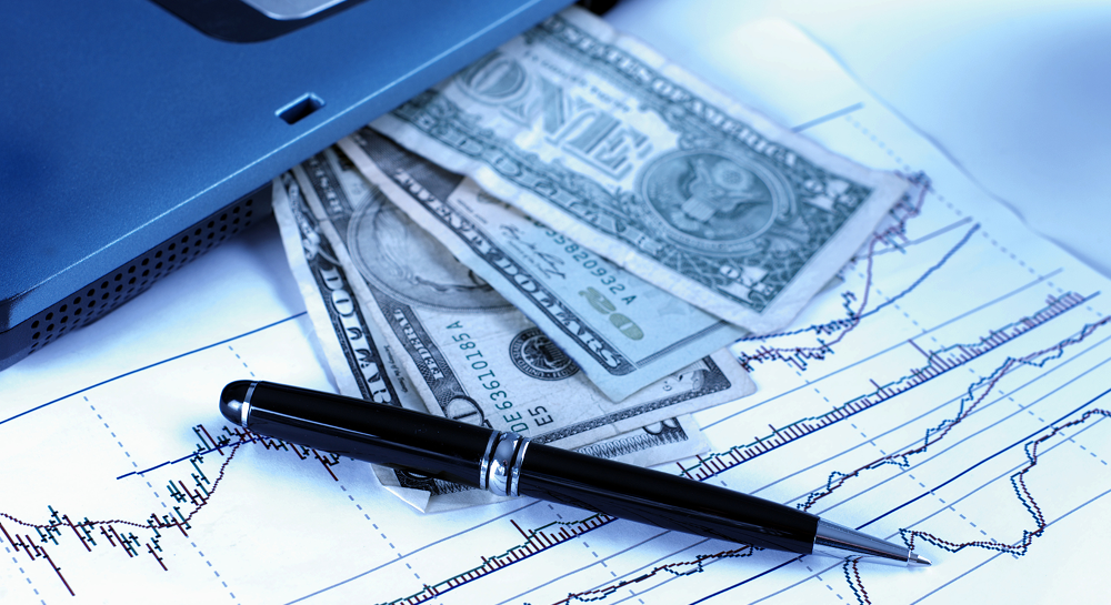 US dollar bills, a pen, and financial charts beside a laptop.