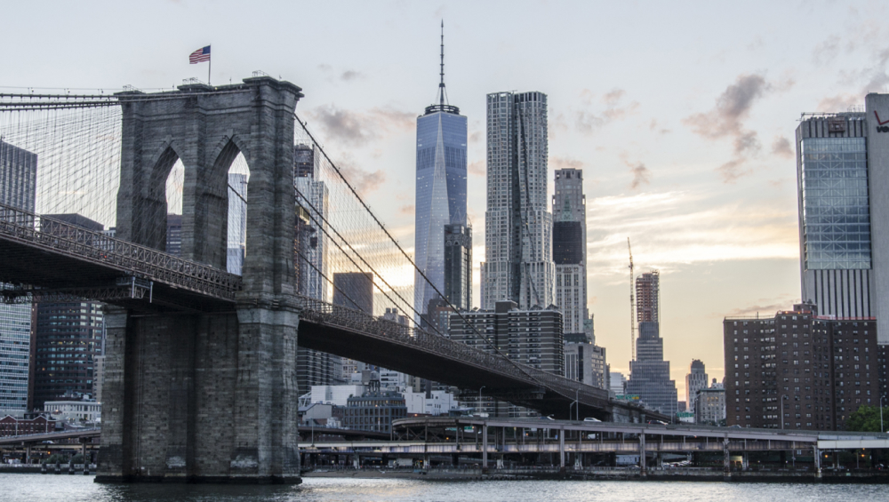 The Brooklyn Bridge's stone arches and cables in the foreground, with the modern One World Trade Center and surrounding Manhattan skyscrapers in the background at dusk.