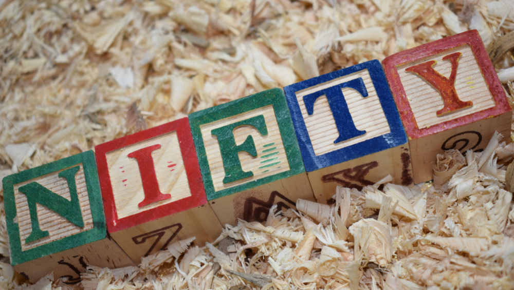 Wooden alphabet blocks spelling the word "NIFTY" laid diagonally on a bed of tan wood shavings or sawdust.