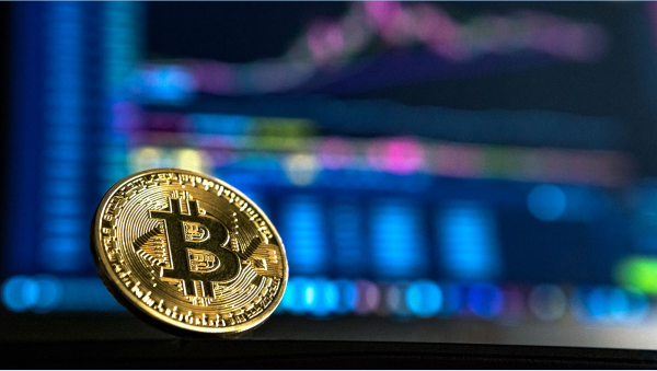 A physical gold-colored Bitcoin coin standing upright against a blurred, glowing blue and pink digital cryptocurrency trading chart background.