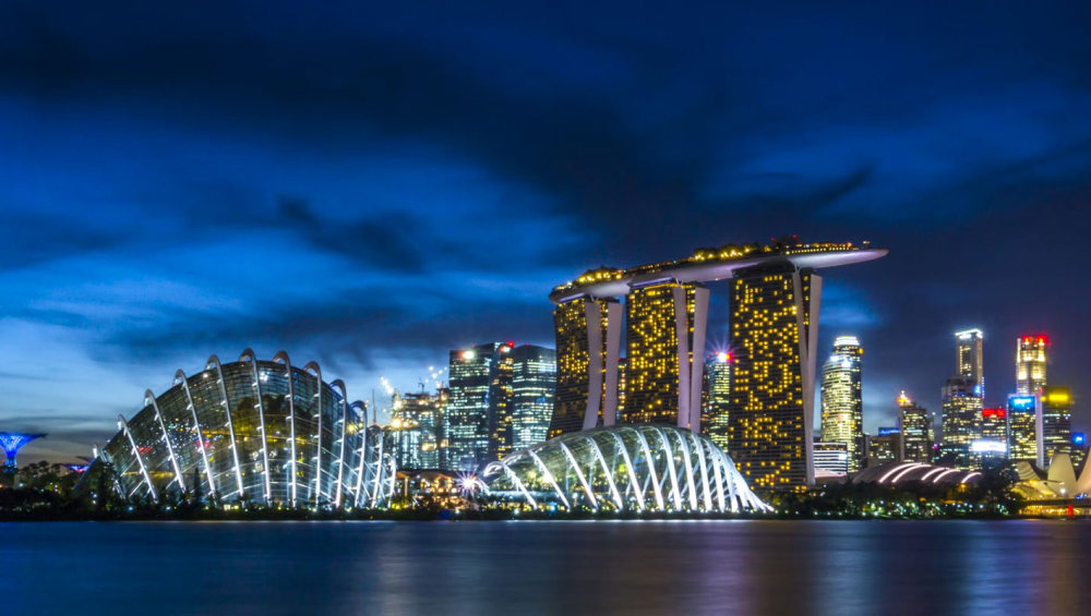 The illuminated skyline of Singapore at night, featuring the Marina Bay Sands hotel and the domes of Gardens by the Bay reflected in the water.