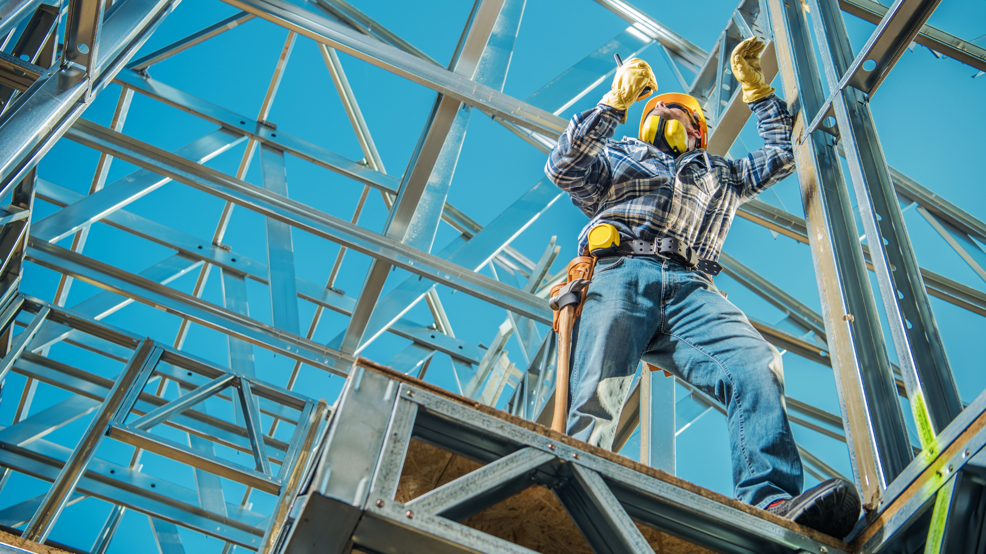 Construction worker in safety gear standing on a steel building frame against a blue sky.