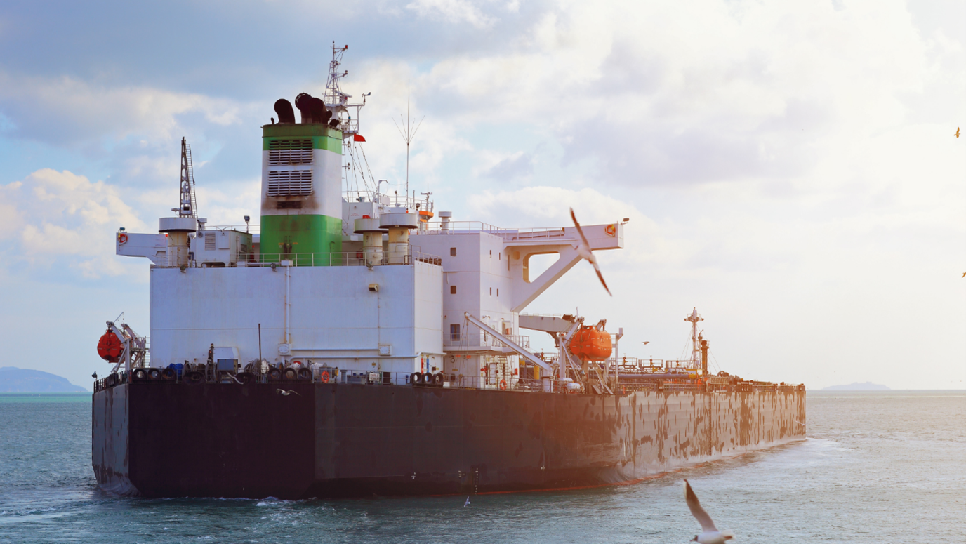 A large, dark hull oil tanker or bulk cargo ship moving on the ocean with its white and green superstructure visible against a bright, cloudy sky.