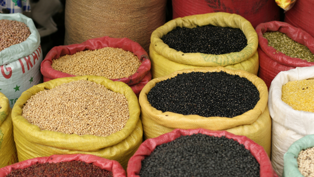 A vibrant display of open burlap and plastic sacks filled with various types of dry grains, pulses, and beans, including black and beige varieties.