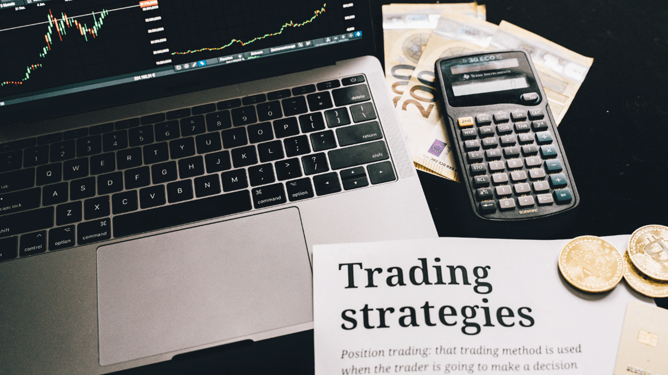 Laptop displaying stock charts, calculator, Euros, Bitcoin, and a "Trading strategies" document.