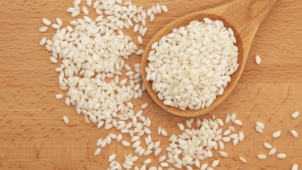 Uncooked white rice grains spilled from a wooden spoon onto a wooden board.