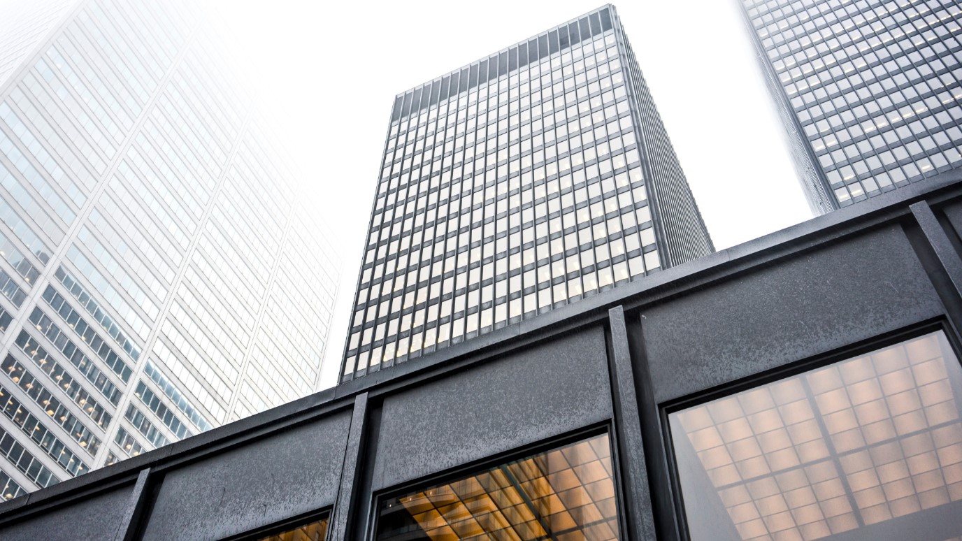 Low-angle view of tall, dark, modern skyscrapers rising above a pedestrian walkway structure on a hazy or foggy day.