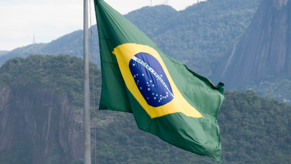 The vibrant green, yellow, and blue Brazilian flag, waving against a backdrop of steep, misty green mountains