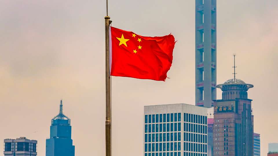 The national flag of China waving on a flagpole against a backdrop of modern skyscrapers in a city.