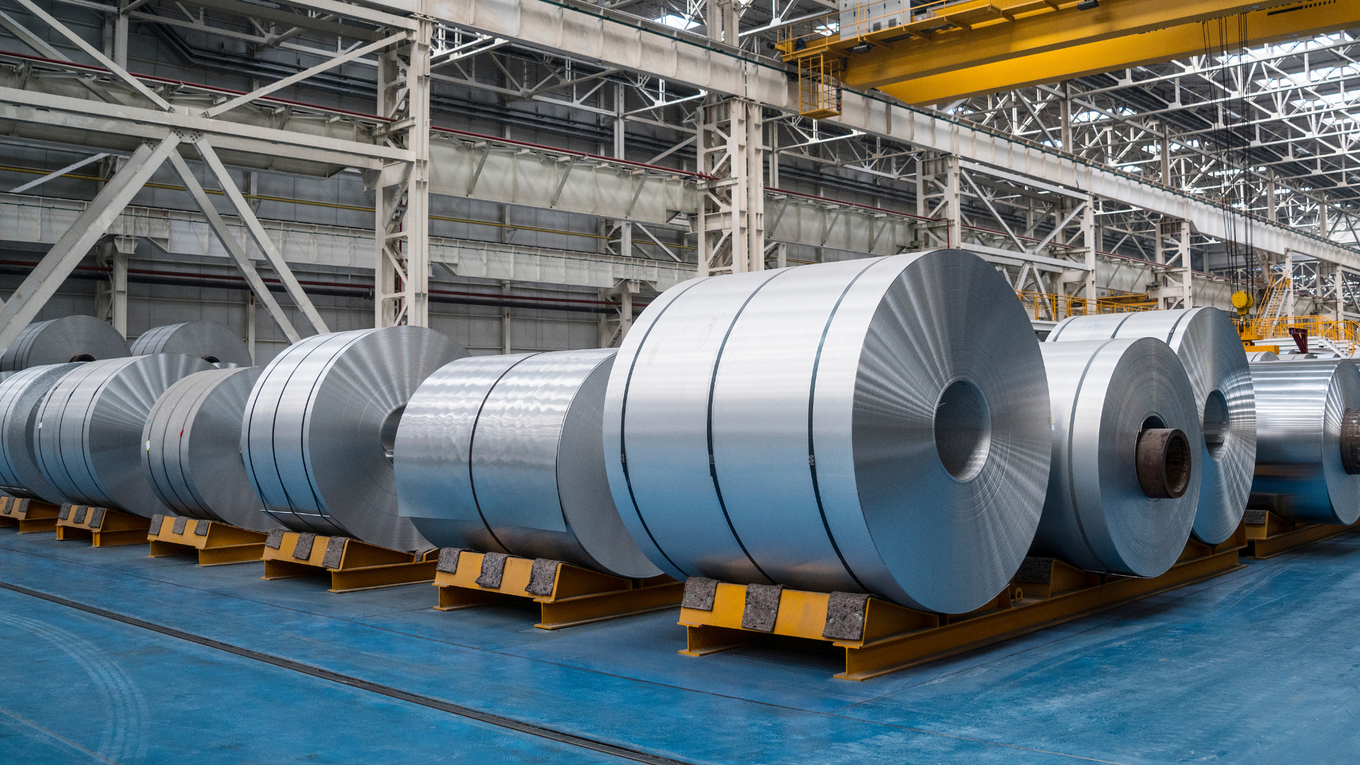 Several large, bright metallic aluminum coils stored horizontally on yellow industrial racks in a factory.