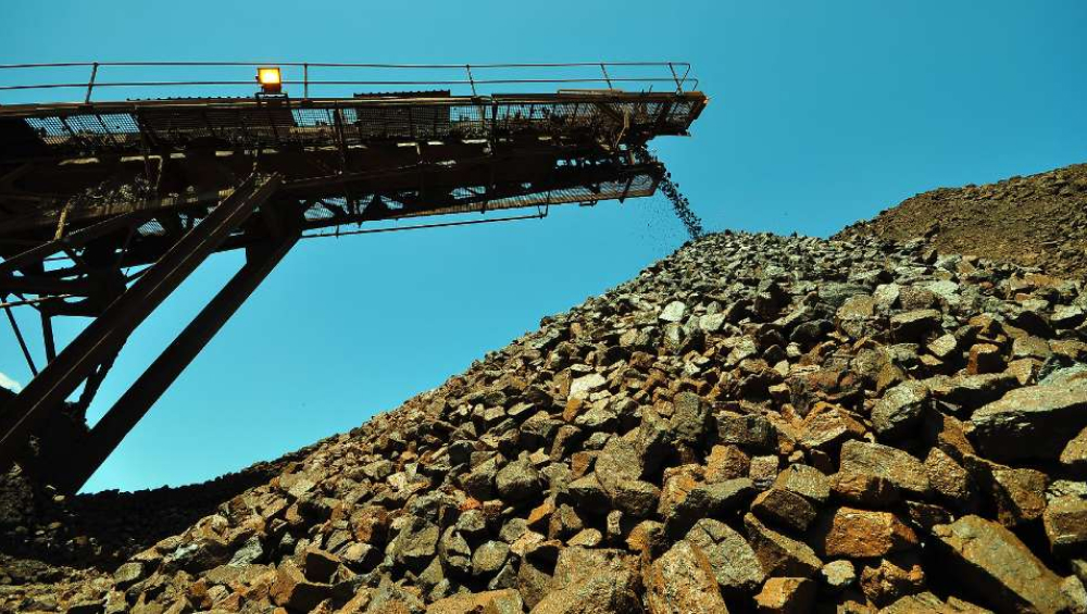 Conveyor belt dropping dark red iron ore rocks onto a massive stockpile under a blue sky.