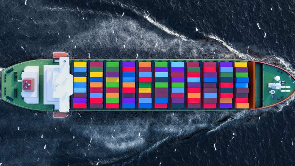 Top-down view of a massive container ship loaded with brightly colored shipping containers sailing on dark blue open water.