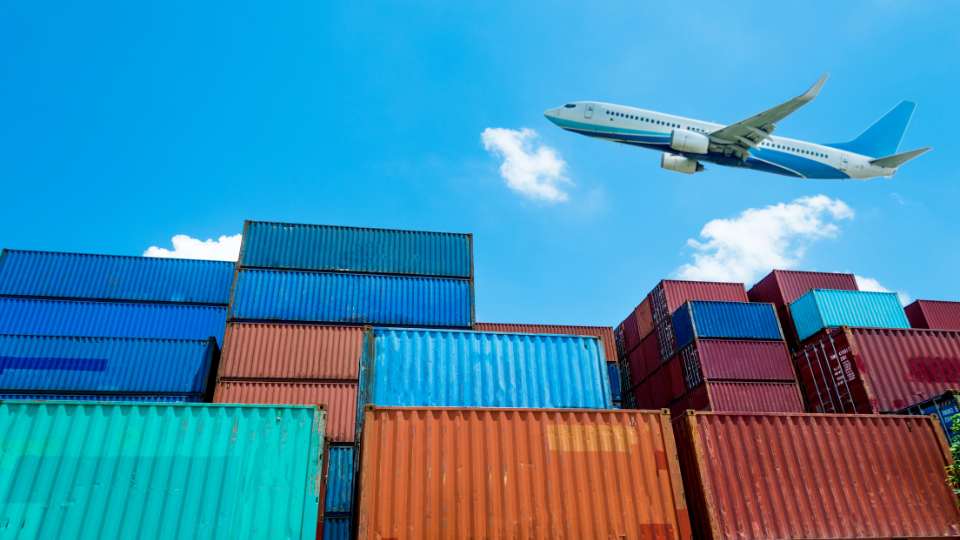 A low-angle view of multi-colored shipping containers stacked at a port terminal, with a white airplane flying overhead against a blue sky.
