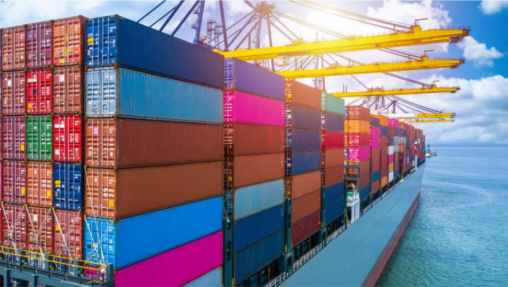 A massive cargo ship loaded with tall stacks of multicolored shipping containers, docked beneath large yellow port cranes under a sunny sky.