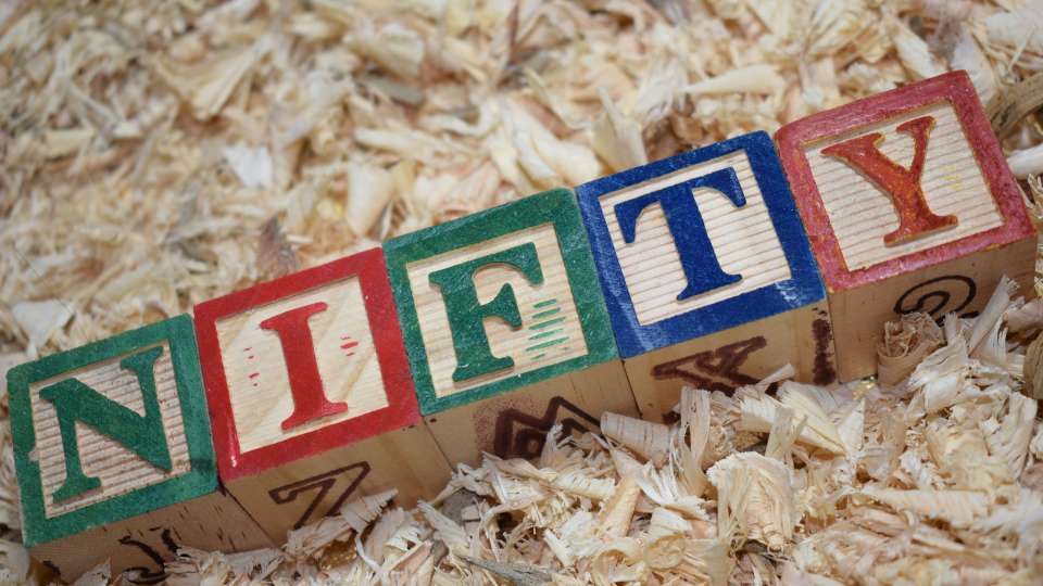 Wooden alphabet blocks spelling the word "NIFTY" laid diagonally on a bed of tan wood shavings or sawdust.