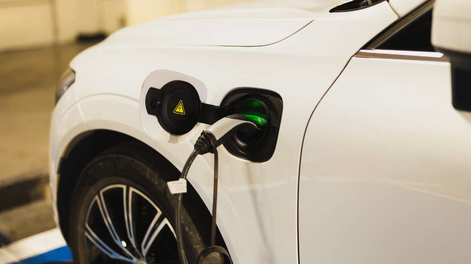 A white electric vehicle (EV) being charged, with the charging plug inserted into the car's port, showing a green indicator light.