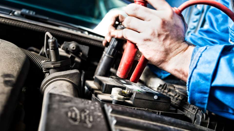 A mechanic's hands connecting a red jumper cable clamp to the terminal of a traditional car battery under the hood of a vehicle.