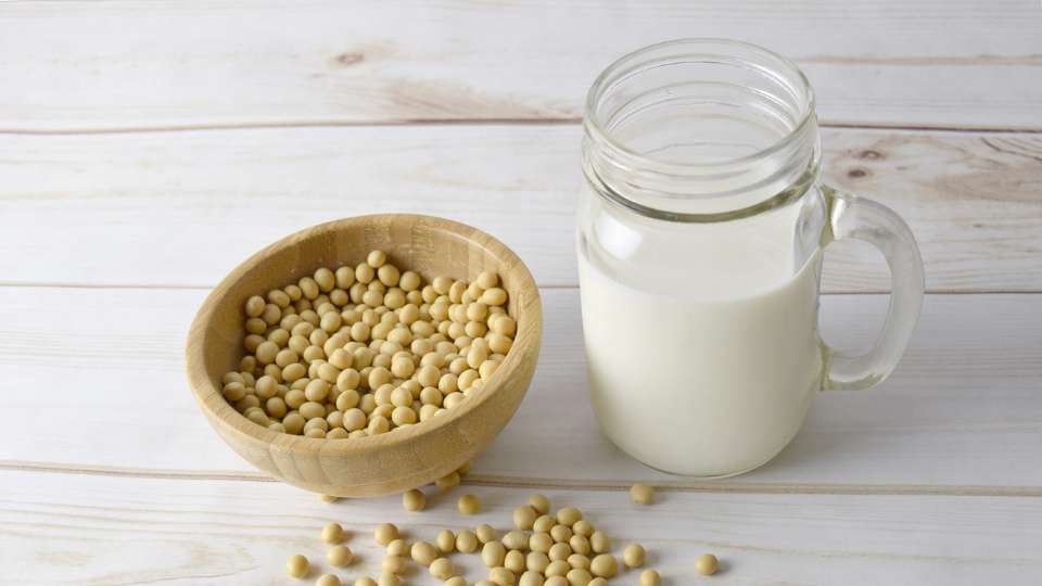 A glass jar mug of soy milk placed next to a small wooden bowl filled with soybeans on a white wooden table.