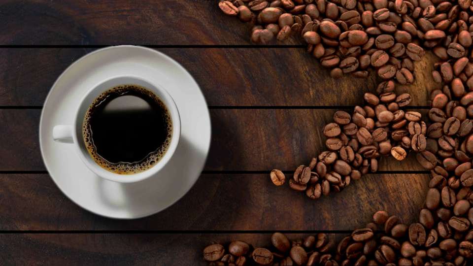 White cup of coffee next to scattered coffee beans on wooden table.