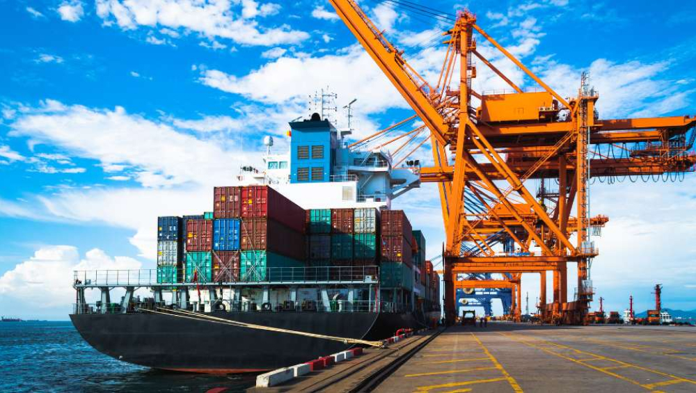 A massive cargo ship loaded with tall stacks of multicolored shipping containers, docked beneath large yellow port cranes under a sunny sky.