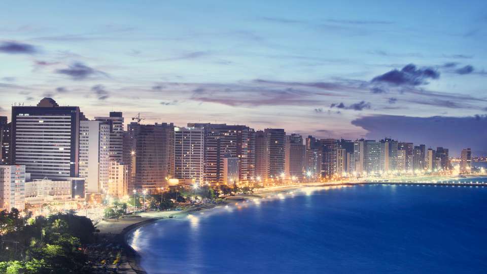 High-angle panoramic view of a densely developed coastal city skyline along a curved beach at dusk in Brazil.