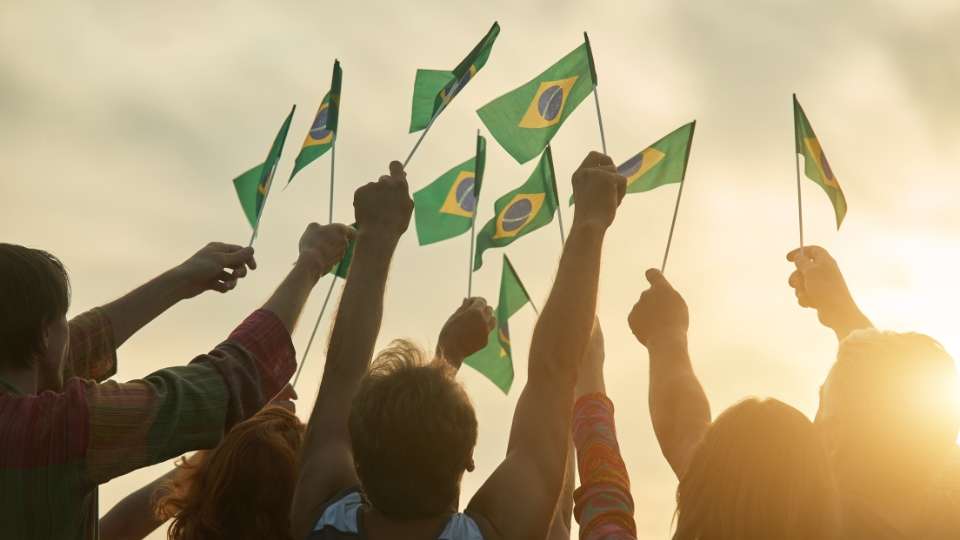 Crowd of people holding Brazilian flags up in the air, silhouetted against a bright, sunny sky.