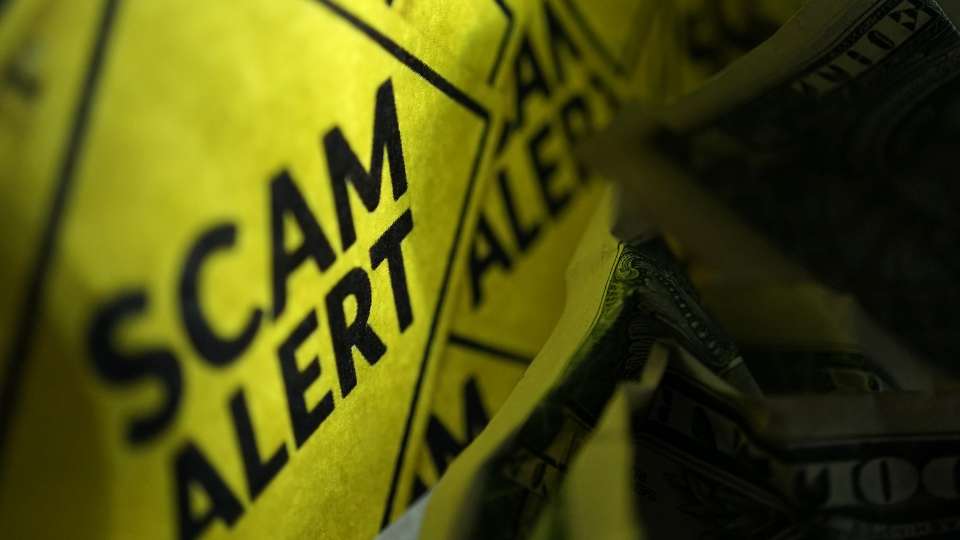 Close-up of a bright yellow sign with the text "SCAM ALERT" in black, partially covering crumpled US dollar bills.