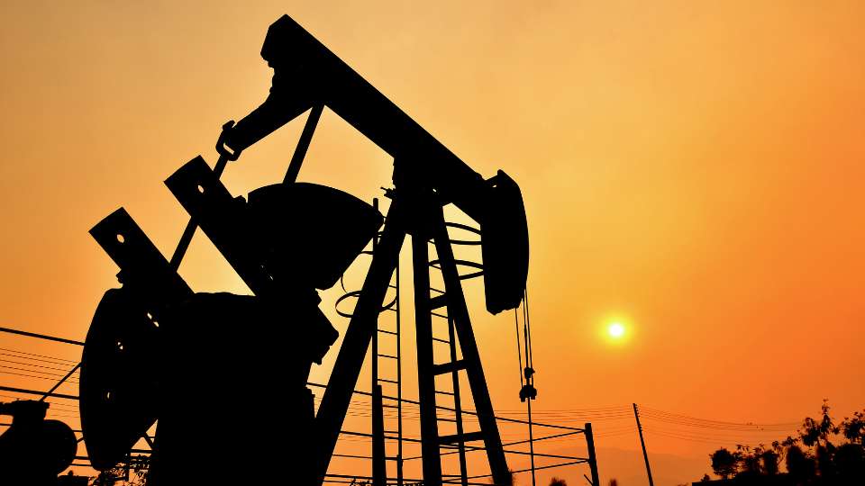 Silhouette of an oil pumpjack operating against a fiery orange sunset or sunrise sky.