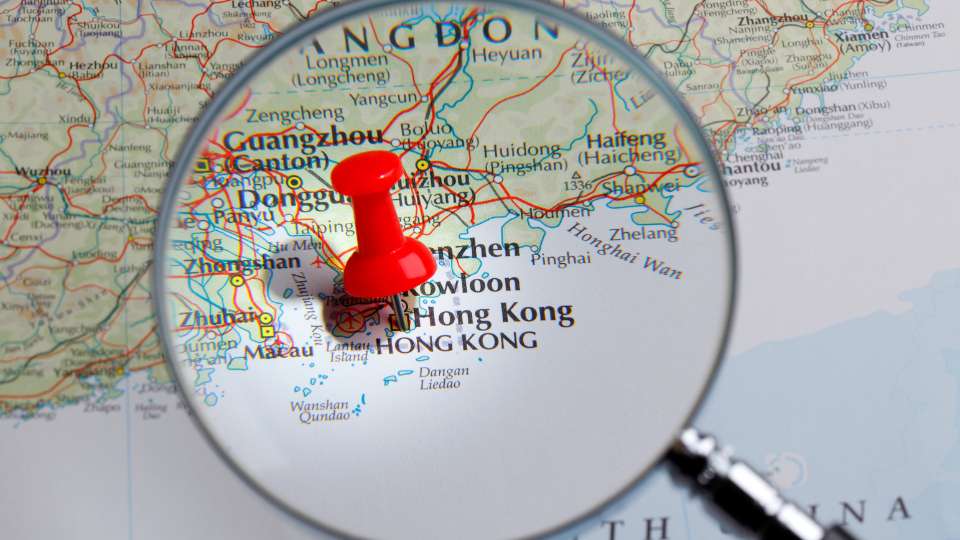 A magnifying glass over a map of southern China, with a red pushpin marking the location of Hong Kong and Shenzhen.