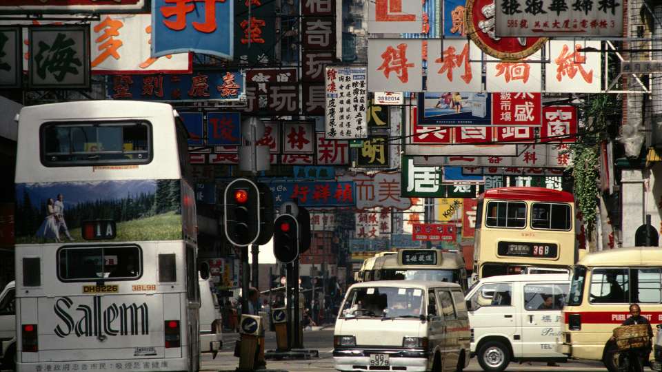Busy street scene in Hong Kong with dense, towering traditional Chinese signs and various buses and vans.