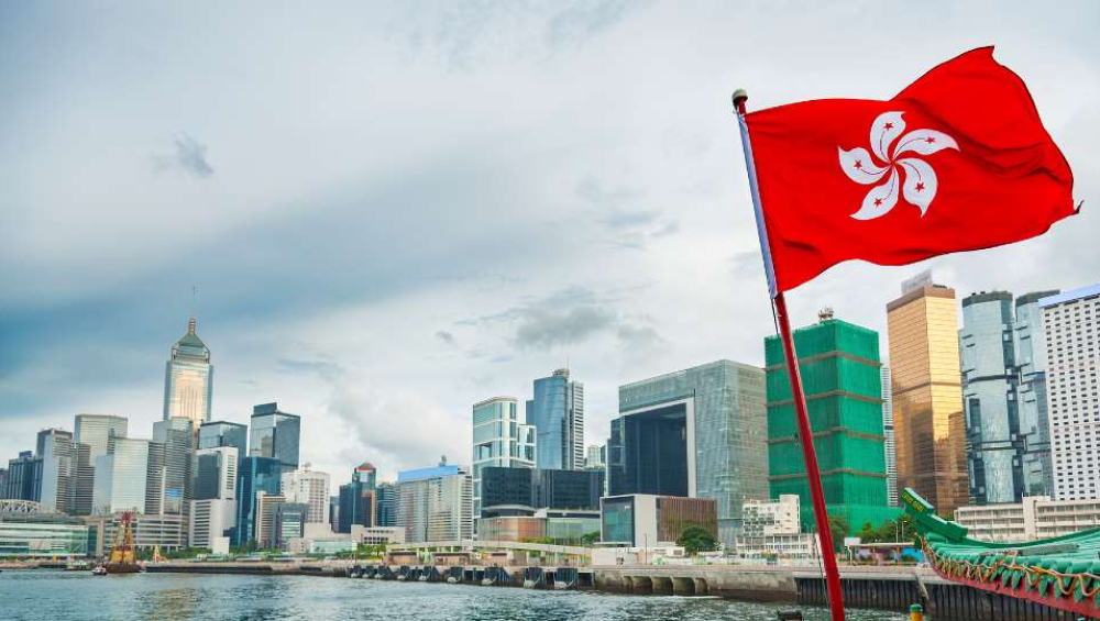 The red flag of Hong Kong waving over the Victoria Harbour skyline under a cloudy sky.